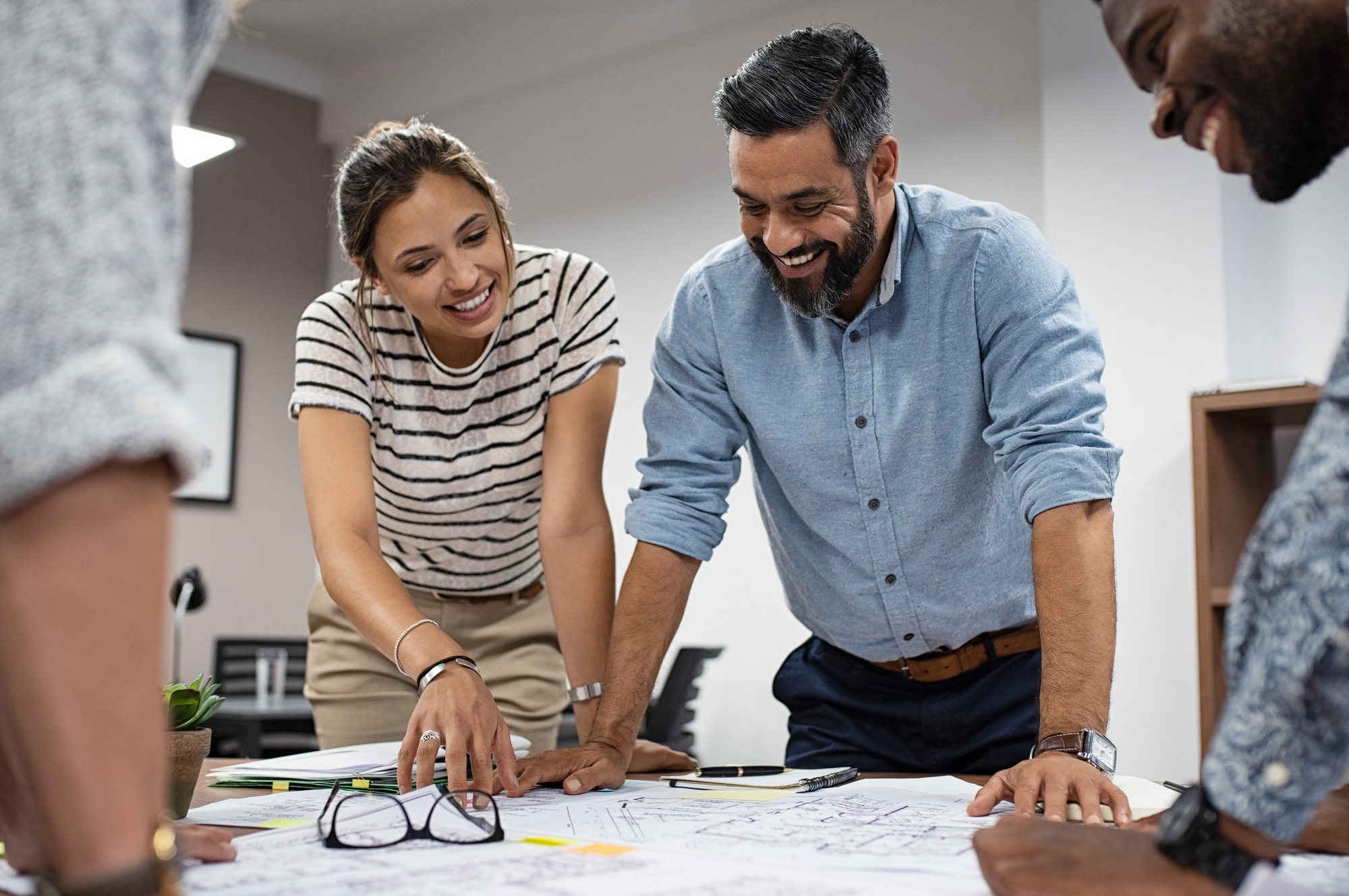 A diverse team collaborates over blueprints on a table, discussing plans with smiles in a modern office setting, promoting teamwork and creativity.